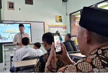 A man in a beige uniform speaks at the front of a classroom or meeting room, addressing students seated at desks while a person in the foreground records with a smartphone.
